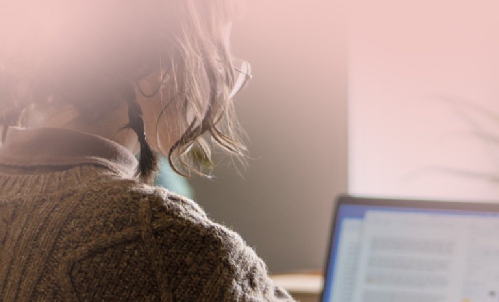 A person with shoulder-length hair, wearing glasses and a knitted sweater, is focused on typing at a laptop. The image has a warm, soft pink overlay.