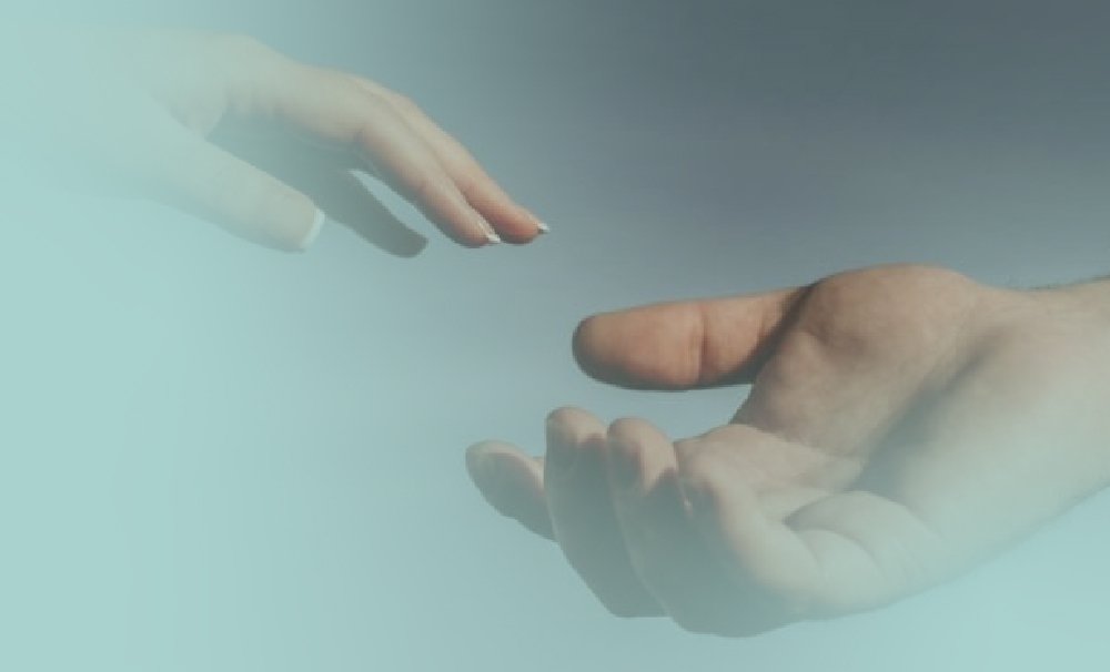 Two hands reaching toward each other against a soft blue background; one hand reaches down from the upper left, while the other reaches up from the lower right, creating a sense of connection and closeness.
