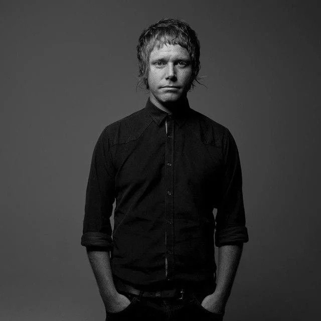 Black and white portrait of a man in a buttoned shirt with hands in pockets, neutral expression, medium length hair, dark background.
