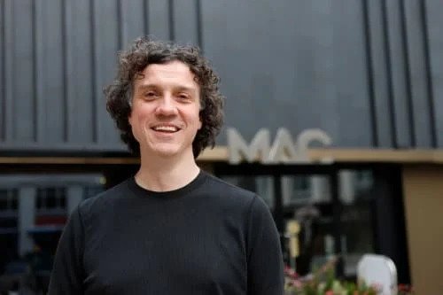 Person with curly hair smiling in front of a building with 'MAC' sign.