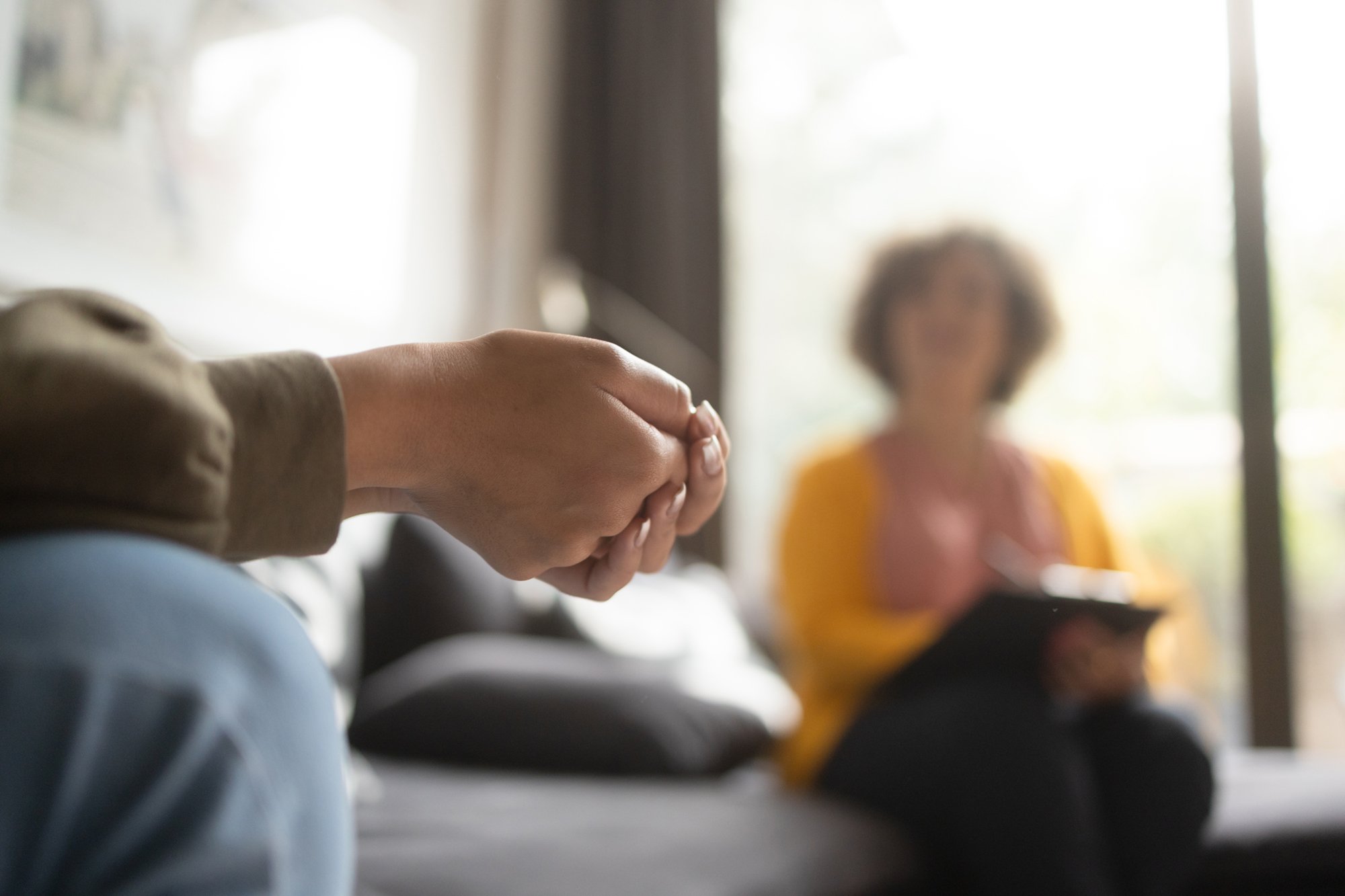 Close-up of a person's hand during a therapy session, with a blurred therapist holding a clipboard in the background.