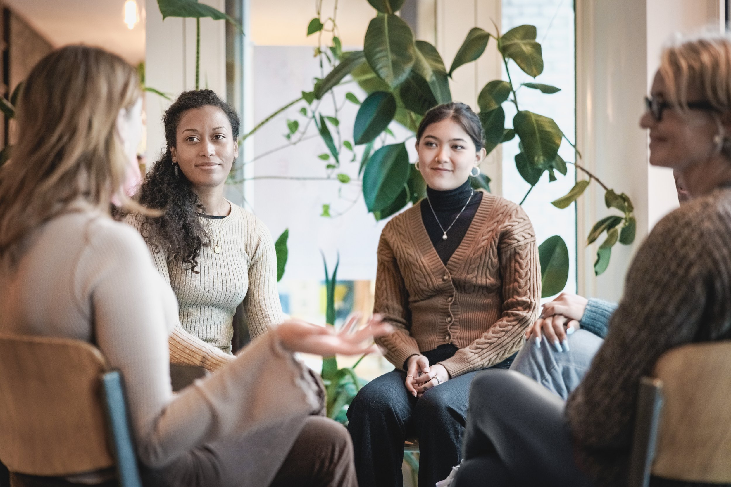 Group of five women sitting in a circle, engaged in conversation, with plants in the background.