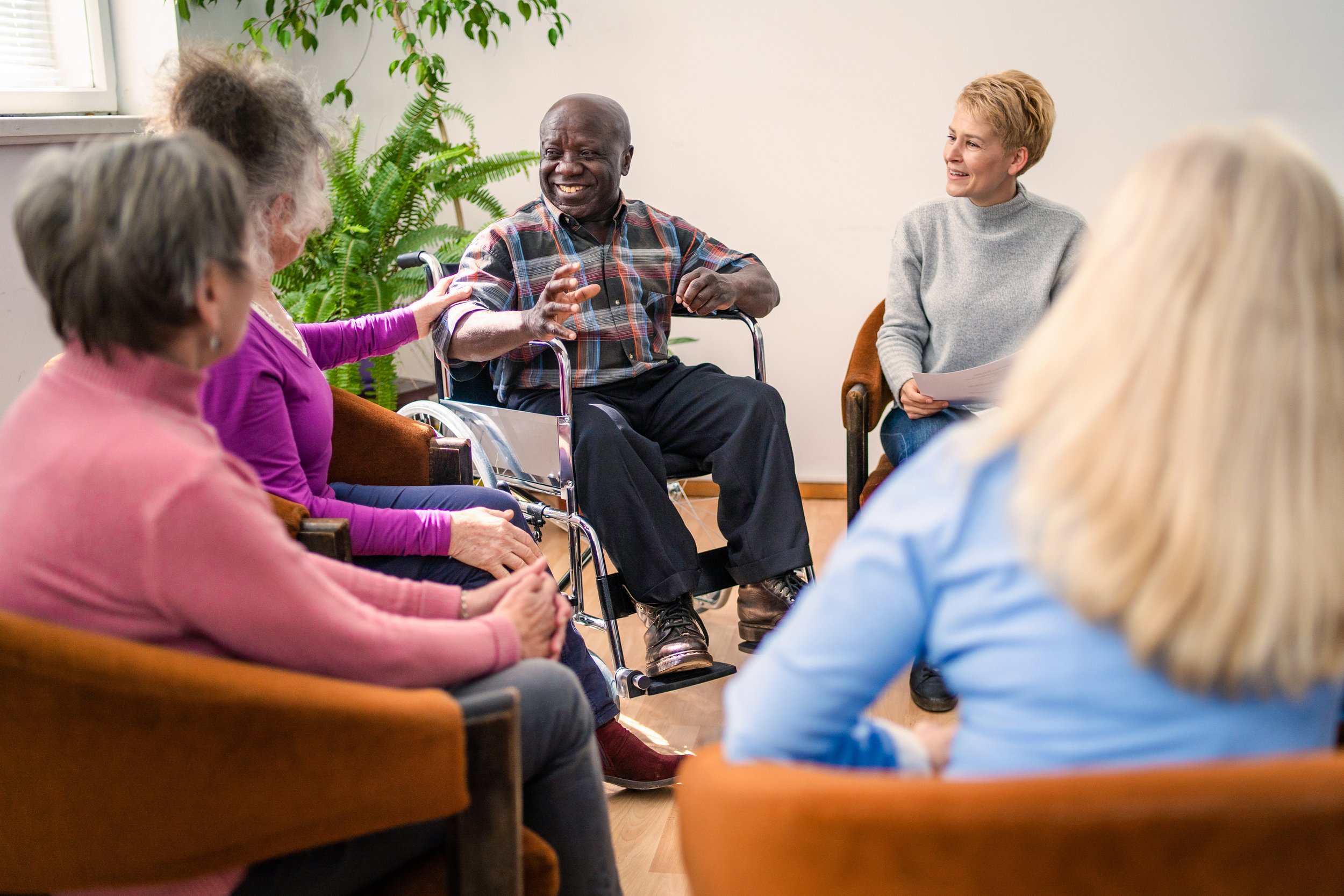 A diverse group of older adults sitting in a circle, engaging in a discussion. One person is in a wheelchair, speaking animatedly. Others are listening and smiling, with plants and a window in the background.