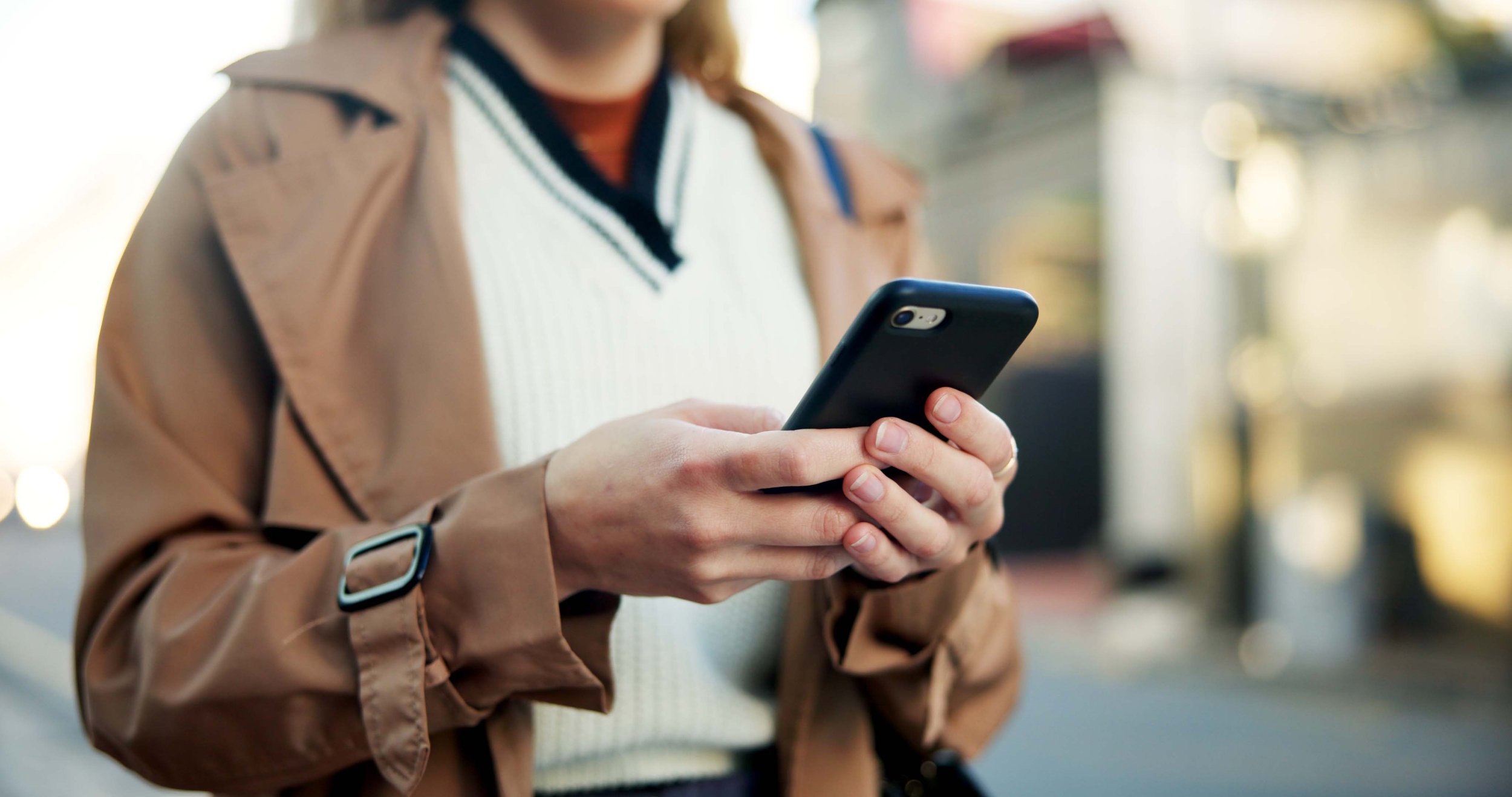 Person holding a smartphone while wearing a brown coat and white sweater.