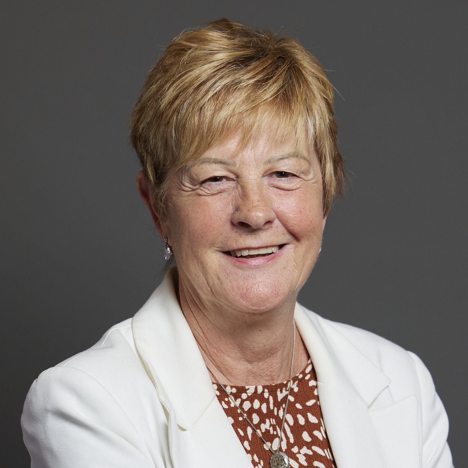 Smiling person wearing a white blazer and patterned top, against a gray background.