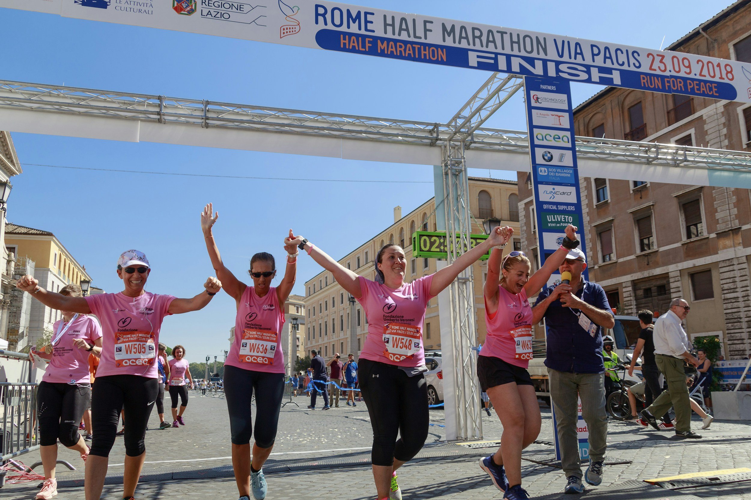 A group of women in pink shirts joyfully crossing a marathon finish line, celebrating their achievement together.