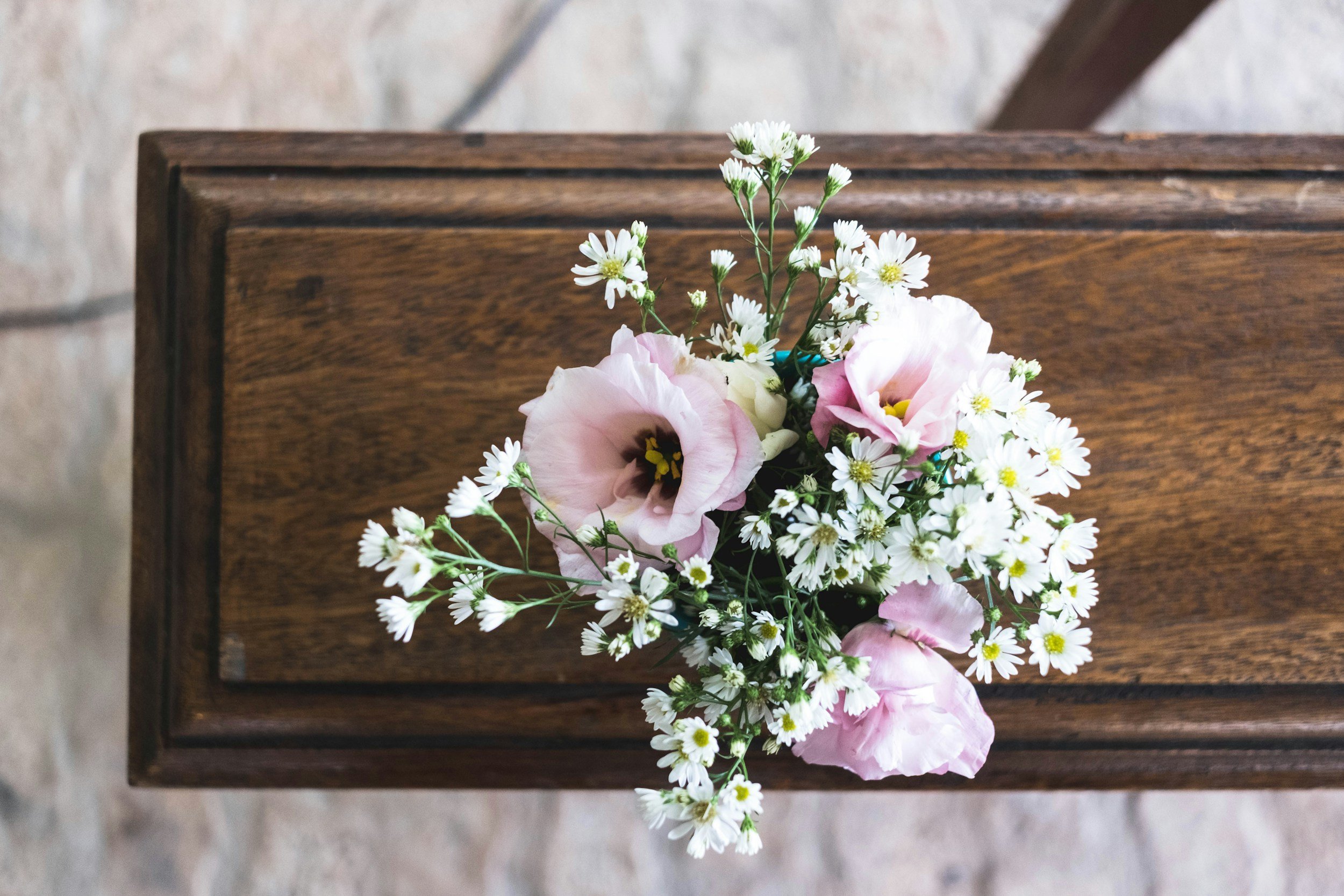 A elegant wooden casket adorned with pink and white flowers rests over a stone floor.
